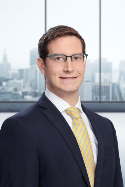 A man in a black suit with a yellow tie and glasses smiles